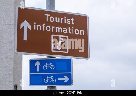 Panneau d'information touristique marron avec lettrage blanc au-dessus des panneaux bleus des pistes cyclables placés contre un ciel nuageux lumineux. Ballycastle, Royaume-Uni - 24 août 2024. Banque D'Images