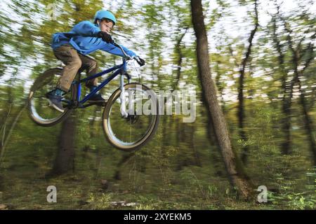 Un jeune cavalier dans un casque et un sweat-shirt bleu vole sur un vélo après avoir sauté d'un haut kicker sur une piste cyclable forestière. Prise de vue avec exposition prolongée Banque D'Images