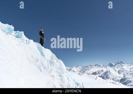 L'alpiniste se tient au bord du glacier avec une pelle à neige dans ses mains et montre le geste de Shak contre le ciel bleu et le mont du Caucase Banque D'Images