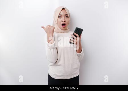 Femme musulmane asiatique choquée portant le hijab pointant vers l'espace de copie à côté d'elle tout en tenant son téléphone, isolée par fond blanc. Banque D'Images