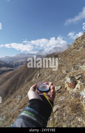 La main d'un homme tient une boussole magnétique de poche pour la navigation sur le fond d'une pente rocheuse et des rochers épiques sous un ciel bleu et des nuages blancs. Banque D'Images