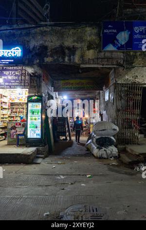 Dhaka, Bangladesh - 05.16.2023, rue nocturne avec un passage étroit éclairé par des panneaux au néon. Une personne se promène le long de magasins entourés de marchandises. Vie nocturne urbaine Banque D'Images