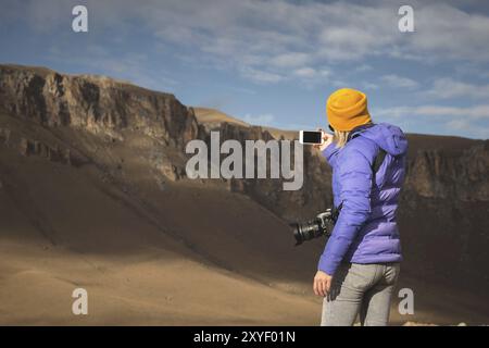 Portrait du dos d'une fille voyageant dans une veste en duvet avec une casquette prenant des photos d'un paysage épique avec des rochers sur son smartphone Banque D'Images