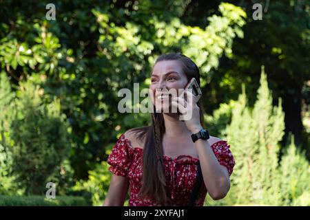 Une jeune fille rousse, avec un sourire insouciant, discute sur son téléphone alors qu'elle marche dans un jardin ensoleillé. Son esprit libre et son énergie positive Banque D'Images