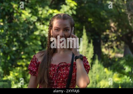 Une jeune rousse aime une promenade insouciante dans un jardin plein de vie, discutant sur son téléphone. La lumière du soleil souligne sa sérénité et sa joie. Banque D'Images