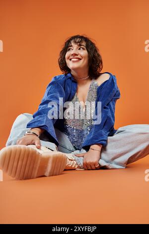 Femme en chemise bleue et top brillant se trouve sur fond orange, regardant joyeux Banque D'Images