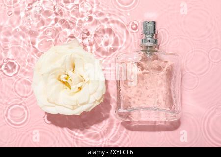 Belle fleur de rose et bouteille de parfum dans l'eau sur fond rose, pose plate Banque D'Images