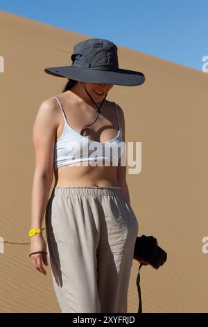 Une femme blanche marchant sur les dunes du désert avec un chapeau et un appareil photo à la main. Banque D'Images