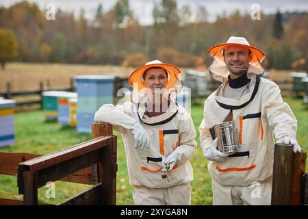 Portrait d'une apicultrice souriante d'âge moyen vêtue d'une combinaison de protection et d'un collègue adulte regardant la caméra tout en se tenant debout à l'entrée de la ferme du rucher à côté de la forêt d'automne, espace copie Banque D'Images