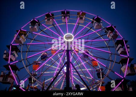 ARLINGTON, Virginie — Une grande roue brillamment éclairée se détache sur le ciel crépusculaire de la foire du comté d'Arlington. Les lumières colorées de la roue créent un spectacle éblouissant, capturant l'atmosphère festive de la foire lorsque le jour se transforme en nuit. Ce carnaval emblématique sert de pièce maîtresse à l'événement communautaire annuel, offrant à la fois des vues palpitantes pour les riders et une scène pittoresque pour les fêtards. Banque D'Images