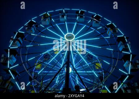 ARLINGTON, Virginie — Une grande roue brillamment éclairée se détache sur le ciel crépusculaire de la foire du comté d'Arlington. Les lumières colorées de la roue créent un spectacle éblouissant, capturant l'atmosphère festive de la foire lorsque le jour se transforme en nuit. Ce carnaval emblématique sert de pièce maîtresse à l'événement communautaire annuel, offrant à la fois des vues palpitantes pour les riders et une scène pittoresque pour les fêtards. Banque D'Images