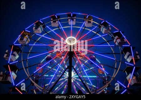 ARLINGTON, Virginie — Une grande roue brillamment éclairée se détache sur le ciel crépusculaire de la foire du comté d'Arlington. Les lumières colorées de la roue créent un spectacle éblouissant, capturant l'atmosphère festive de la foire lorsque le jour se transforme en nuit. Ce carnaval emblématique sert de pièce maîtresse à l'événement communautaire annuel, offrant à la fois des vues palpitantes pour les riders et une scène pittoresque pour les fêtards. Banque D'Images