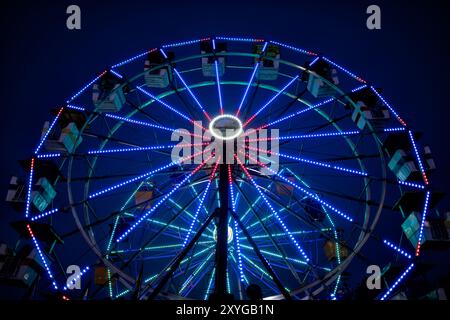 ARLINGTON, Virginie — Une grande roue brillamment éclairée se détache sur le ciel crépusculaire de la foire du comté d'Arlington. Les lumières colorées de la roue créent un spectacle éblouissant, capturant l'atmosphère festive de la foire lorsque le jour se transforme en nuit. Ce carnaval emblématique sert de pièce maîtresse à l'événement communautaire annuel, offrant à la fois des vues palpitantes pour les riders et une scène pittoresque pour les fêtards. Banque D'Images