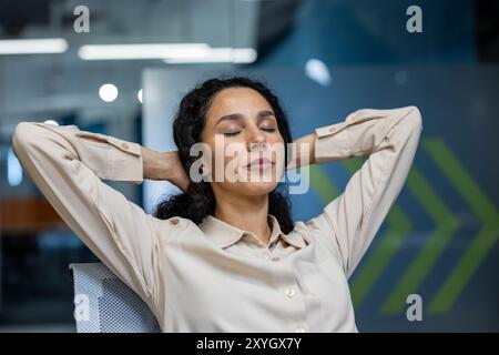 Femme d'affaires confiante au bureau prend un moment pour se détendre avec les yeux fermés et les mains derrière la tête. Portant une chemise blanche élégante, elle incarne le calme et la tranquillité pendant les pauses au travail. Banque D'Images