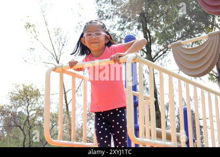 Fille brune Latina de 4 ans avec des lunettes joue sur le toboggan de l'aire de jeux dans le parc profite du week-end de vacances Banque D'Images