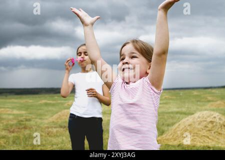 Petite fille gaie attrapant des bulles de savon tout en s'amusant avec une sœur plus âgée dans le champ vert en été avec ciel nuageux orageux en arrière-plan Banque D'Images