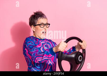 Photo de la femme senior attrayante effrayée accident tenir le volant habillé de vêtements colorés bleus isolés sur fond de couleur rose Banque D'Images