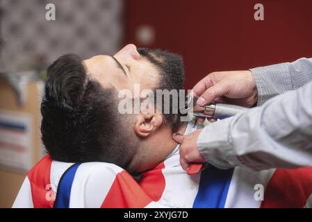 Barbier rasant la barbe d'un bel homme barbu avec un rasoir électrique au salon de coiffure Banque D'Images