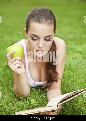 Belle jeune fille allongée sur l'herbe et lisant dans le parc d'été Banque D'Images