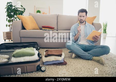 Photo d'un bel homme d'âge mûr assis à l'intérieur du salon emballant des vêtements en utilisant une liste spéciale pour sa tournée à l'étranger préparant une valise Banque D'Images