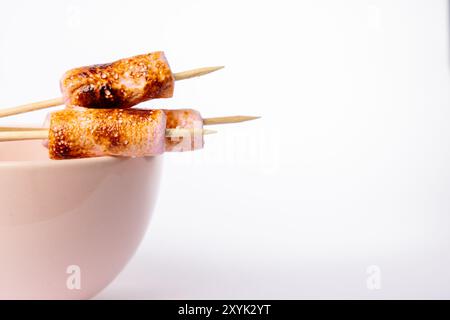 Gros plan de brochettes avec des guimauves grillées dans un bol rose, disposées à gauche sur un fond blanc. La photo met en évidence le brun doré Banque D'Images