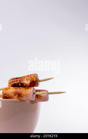 Plan vertical détaillé de brochettes de guimauve, grillées à la perfection, dans un bol rose sur fond blanc. Les brochettes sont disposées sur la gauche, en haut Banque D'Images
