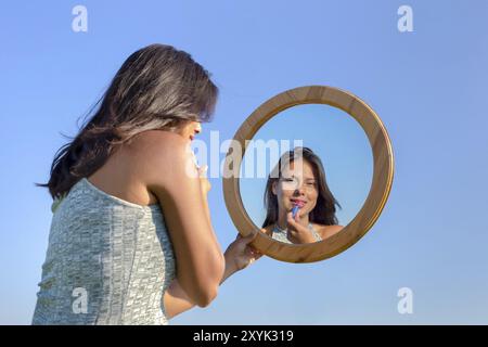 Jolie femme d'appliquer des cosmétiques rouge à lèvres à l'extérieur de miroir avec ciel bleu Banque D'Images