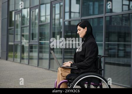 Une femme d'affaires en fauteuil roulant utilise un ordinateur portable lorsqu'elle est assise à l'extérieur d'un immeuble de bureaux. Banque D'Images