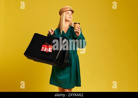 Jeune femme élégante en trench-coat vert portant des sacs à provisions noirs et buvant du café sur fond de studio jaune Banque D'Images