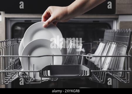 Une femme a fait la vaisselle à la main, a vidé ou a déchargé le lave-vaisselle avec les ustensiles. Appareils de cuisine, vue sur le style de vie. Une femme met une plaque au lave-vaisselle Banque D'Images