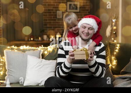 Joyeux Noël et Bonne Année. Surprise cadeau de Noël. Une petite fille souriante embrassant le père dos tenant le cadeau à l'intérieur à la maison décoré Banque D'Images