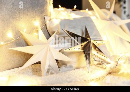 Décorations d'ambiance de Noël ou de Happy Time du nouvel an, forme d'étoile en papier argenté, guirlandes lumineuses électriques de Noël sur un canapé avec oreillers et couverture blancs, Banque D'Images