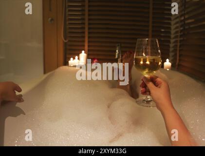 Vue arrière des pieds de la jeune femme à boire le vin blanc dans la baignoire avec de la mousse et des bougies Banque D'Images