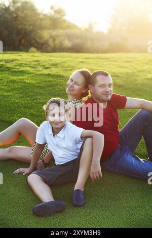 Famille heureuse avec fils assis sur l'herbe dans le parc. Coucher du soleil d'été Banque D'Images