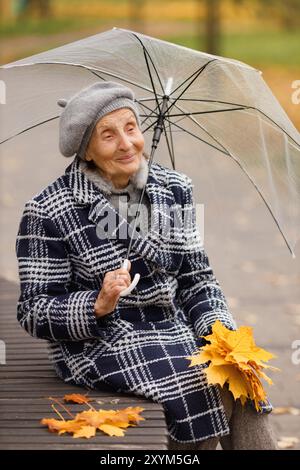 Femme âgée en manteau gris avec parapluie sur le banc avec des feuilles d'automne Banque D'Images