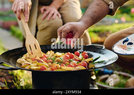 Main de fermier mâle méconnaissable mettant des tomates cerises fraîches sur une poêle à frire avec des légumes hachés tandis que la femme les mélange Banque D'Images