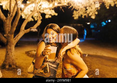 Deux jeunes femmes s'embrassant et partageant un moment joyeux tout en tenant un verre lors d'un événement nocturne en plein air. Banque D'Images