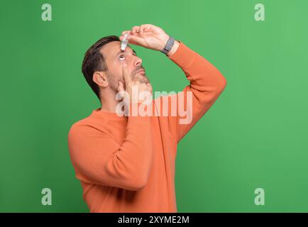 Jeune homme laissant tomber le lubrifiant pour les yeux pour traiter l'oeil sec ou l'allergie tout en se tenant debout sur fond vert Banque D'Images