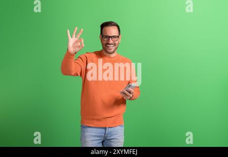 Portrait d'homme souriant positif à l'aide d'un smartphone et montrant le signe OK isolé sur fond vert Banque D'Images