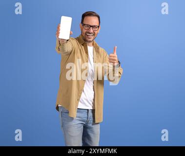 Portrait de jeune homme heureux montrant l'écran blanc du téléphone intelligent et le signe pouces vers le haut sur fond bleu Banque D'Images