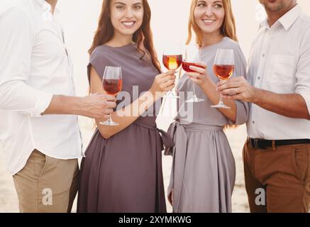 Gai femmes et hommes adultes dans des vêtements décontractés toaster avec des verres à vin reposant ensemble sur la rive et pique-nique au coucher du soleil d'été Banque D'Images