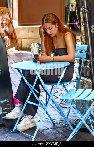 Jeune femme attrayante avec de longs cheveux auburn assise à la table du café regardant smartphone - Tours, Indre-et-Loire (37), France. Banque D'Images