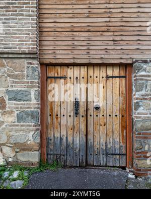 La porte en bois robuste présente des détails complexes en fer, placés contre un mur de pierre altérée, suggérant une histoire riche dans cette région pittoresque. L'herbe luxuriante et les ombres douces rehaussent le charme de la scène Banque D'Images