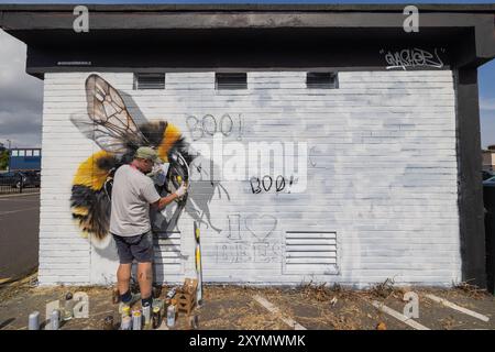 Southend on Sea, Royaume-Uni. 30 août 2024. Un artiste de rue peint activement une grande murale réaliste d'abeilles sur un mur de briques blanches en utilisant de la peinture au pistolet. La murale présente des détails jaunes et noirs vibrants, avec des éléments de graffiti luents, y compris « BOO ! » Et « J’aime les ABEILLES ». Des bombes aérosols sont dispersées autour de l’artiste, capturant le processus créatif et le message environnemental. City Jam ouvre officiellement, avec plus de 200 artistes du monde entier peignant des peintures murales. Penelope Barritt/Alamy Live News Banque D'Images