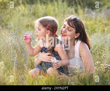 Femme adulte joyeuse avec les yeux fermés souriant et embrassant la petite fille soufflant des bulles de savon tout en étant assis dans l'herbe verte dans la prairie Banque D'Images