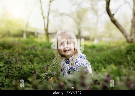 Petite fille souriante avec des fleurs de printemps sur la pelouse verte dans le jardin. Banque D'Images