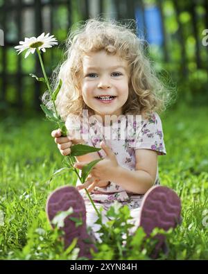 Enfant souriant heureux avec fleur assis sur l'herbe verte à l'extérieur dans le jardin de printemps Banque D'Images