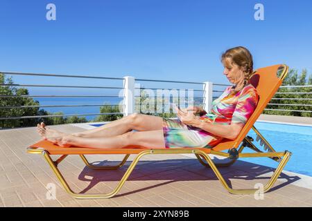 Femme hollandaise d'âge moyen allongée sur une tablette de lecture de transats orange près de la piscine Banque D'Images