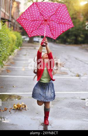 Parapluie enfant à pois rouge portant des bottes de pluie sauter dans une flaque d'eau Banque D'Images