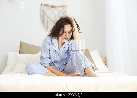 Jeune femme sérieuse en pyjama assis sur le lit le matin Banque D'Images